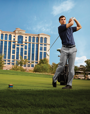 A golfer finishing a swing on a bright green course with the Belterra hotel rising in the background.