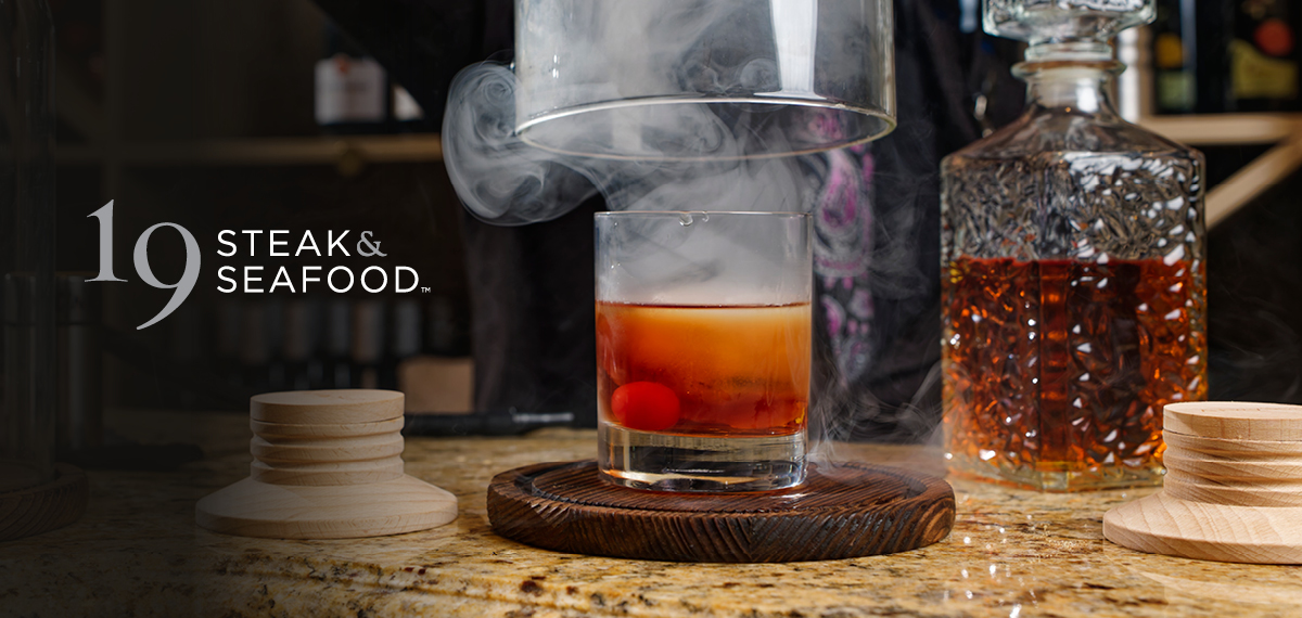 Smoked Old Fashioned cocktail with a cherry garnish under a glass cloche.