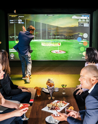 People enjoying conversation over cocktails and snacks while a man swings a golf club in the background