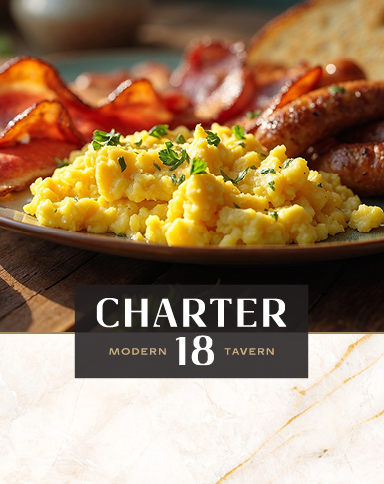 A plate of scrambled eggs, bacon, sausages, and toast on a wooden table with “Charter 18 Modern Tavern” below.