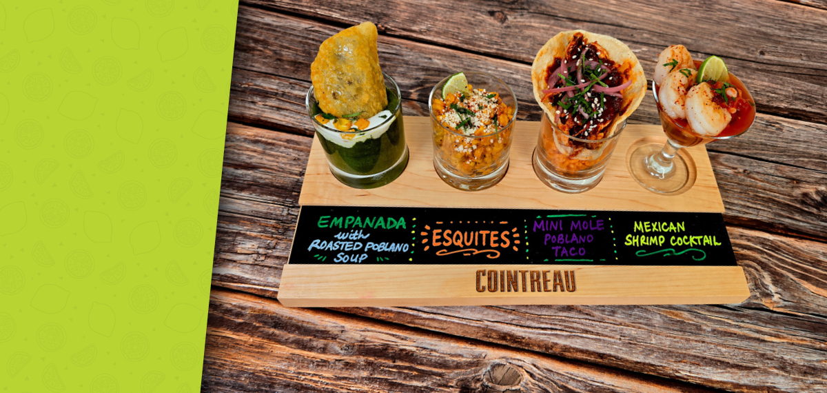 A wooden tasting board holds four small Mexican dishes: an empanada with green poblano soup, a cup of esquites, a mini mole taco, and a shrimp cocktail garnished with lime.