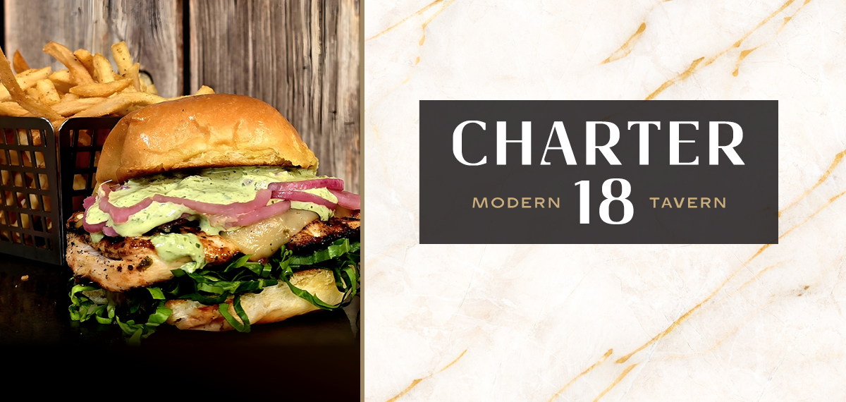 A chicken sandwich with greens and onions beside a basket of fries on a wooden table. Charter 18 Modern Tavern.