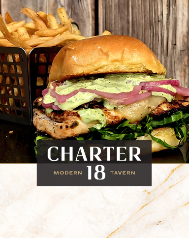 A chicken sandwich with greens and onions beside a basket of fries on a wooden table. Charter 18 Modern Tavern.