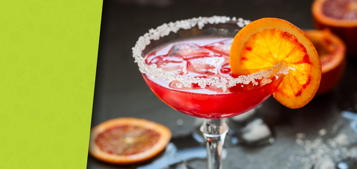 Red citrus cocktail in a salt-rimmed glass with ice and a blood orange garnish, surrounded by ice cubes and orange slices.