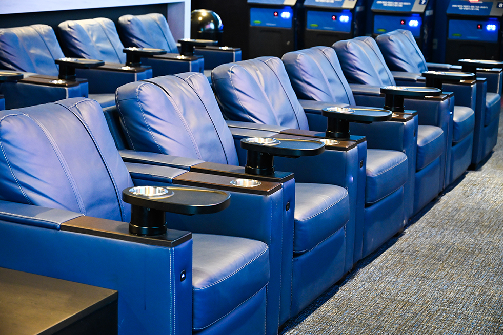 Rows of plush blue leather theater-style seats with built-in cup holders in a sportsbook, arranged in front of multiple television screens.