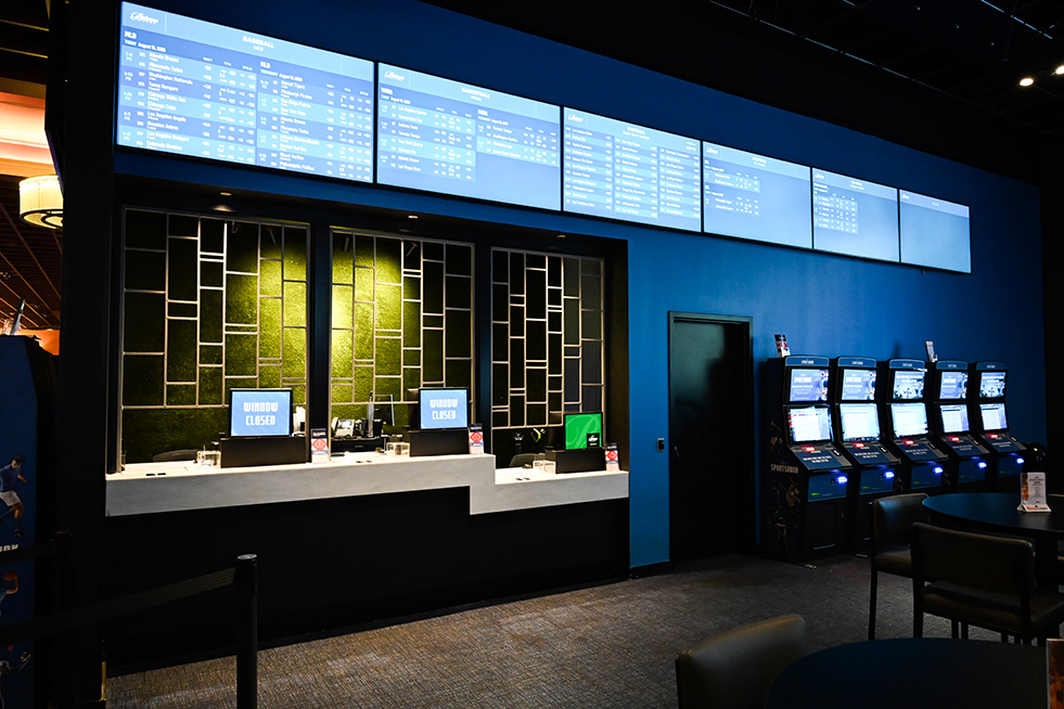 Sportsbook ticket windows beneath large digital betting boards displaying sports odds and wagering information, with self-service betting kiosks and seating area nearby.