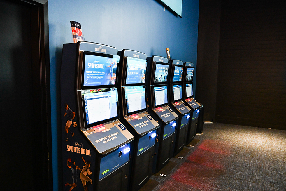 A row of self-service sportsbook betting kiosks with touchscreen displays, allowing guests to place wagers and view sports betting information in a modern sportsbook setting.