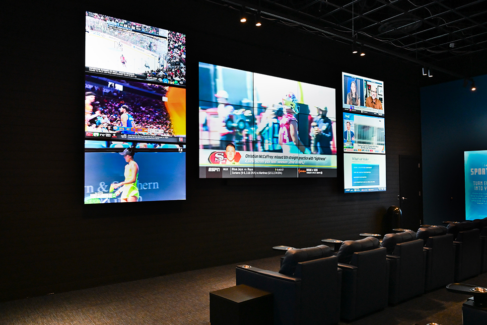 Rows of plush blue leather theater-style seats with built-in cup holders in a sportsbook, arranged in front of multiple television screens.